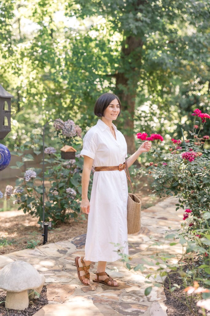 Woman wearing a white cotton midi shirtdress cinched with a braided cognac leather belt, paired with cognac gladiator-style leather sandals and a natural woven straw crossbody bag in a garden with roses and hydrangeas — a polished summer white dress outfit idea.