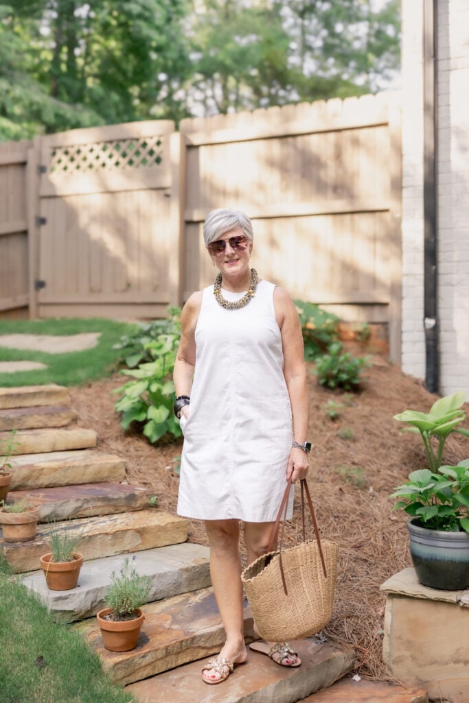 Woman over 50 with silver hair wearing a sleeveless white cotton sheath dress with a sculptural gold and brass statement necklace, aviator sunglasses, embellished gold slide sandals, and a natural straw tote with leather handles on a garden path — a chic minimalist summer white dress outfit for women over 50.