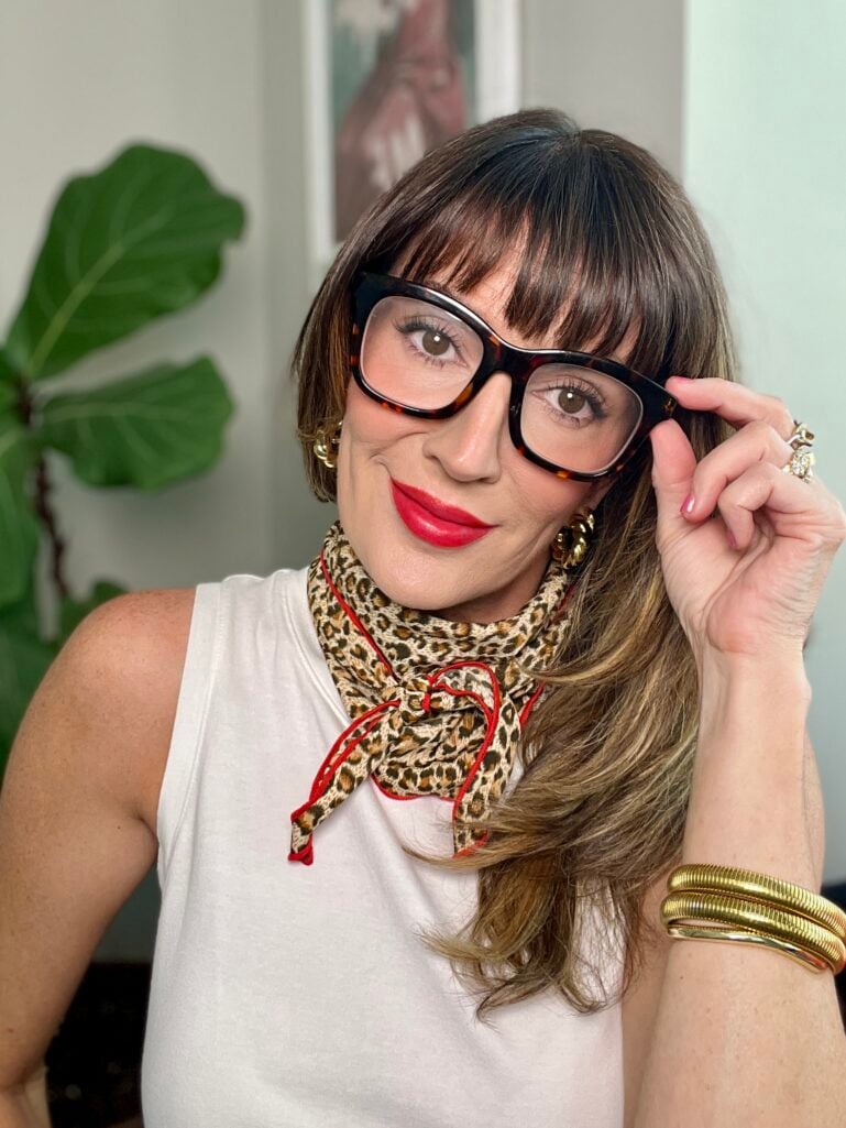 Woman wearing tortoiseshell oversized square glasses, white sleeveless top, leopard print neckerchief with red trim tied in a bow, gold flower earrings and stacked gold tube bangles, showing finished French girl red lip makeup look with diffused matte red lipstick and soft glowy skin, fiddle leaf fig plant in background