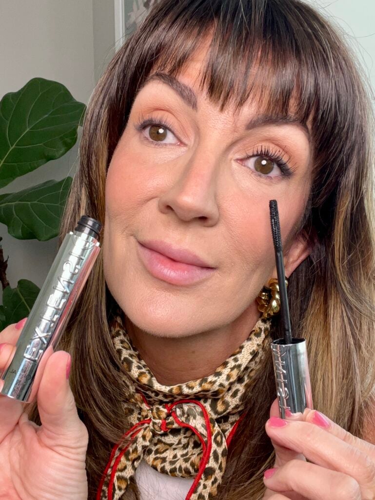 Woman holding L'Oreal Paris Extensionist lengthening and curling mascara wand, showing the dramatic difference between one eye with mascara on full curled lashes and one bare eye without, demonstrating the lash lifting effect, wearing leopard neckerchief and gold flower earrings