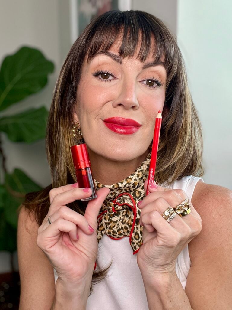 Woman wearing finished French girl red lip holding Clarins Hydrating Matte Water lip stain in Red Water and e.l.f. Cosmetics Glide-On lip liner in Red Receipt, wearing white tank, leopard neckerchief with red trim, gold earrings, and stacked gold rings, fiddle leaf fig in background