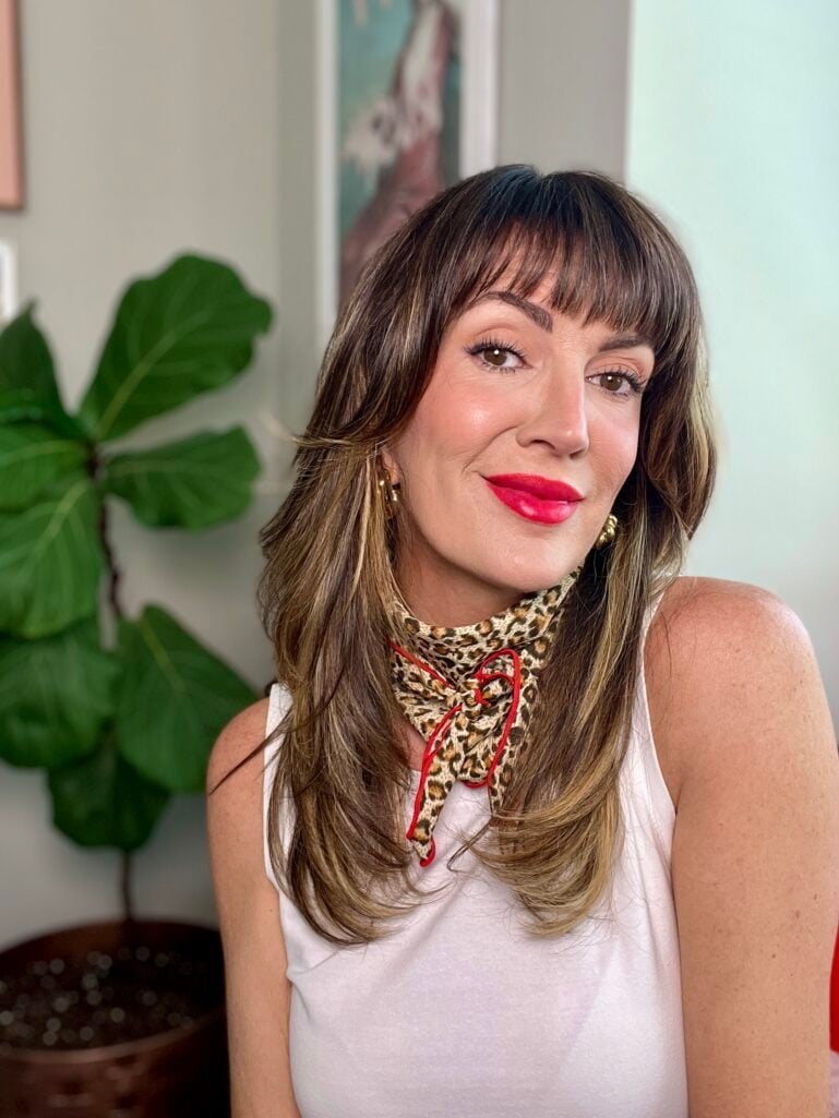 Woman with brown wavy hair and curtain bangs wearing white sleeveless tank top and leopard print neckerchief tied with red trim bow, showing finished French girl summer makeup look with glowy skin, soft brown eyes, fluttery lashes, and diffused matte red lip, fiddle leaf fig plant in background