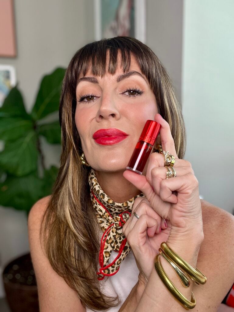 Woman with brown hair and curtain bangs wearing white tank top and leopard print neckerchief with red trim, holding red Clarins Hydrating Matte Water lip stain in Red Water, wearing matching diffused red lip, gold chunky hoop earrings, gold coiled tube bangles, and stacked gold rings