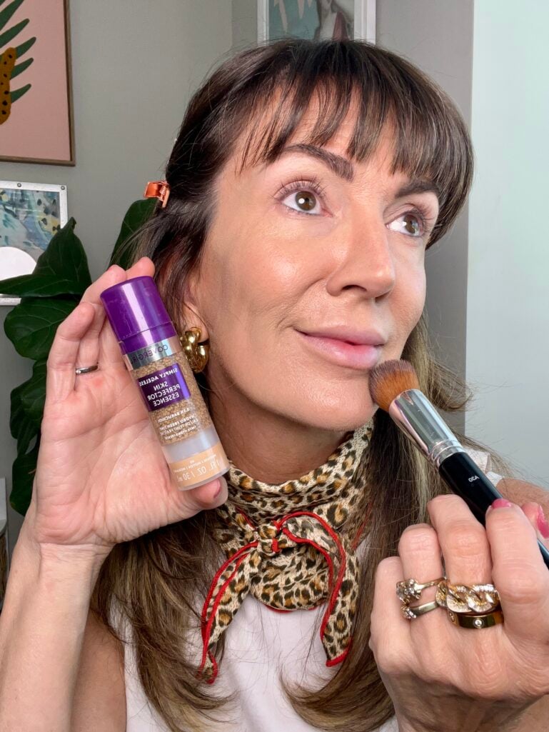 Woman applying CoverGirl Simply Ageless Skin Perfector Essence in shade medium to cheek using gold-handled makeup brush, holding purple foundation bottle, wearing leopard neckerchief with red trim, gold chunky hoop earrings, and stacked gold rings with rose gold hair clip