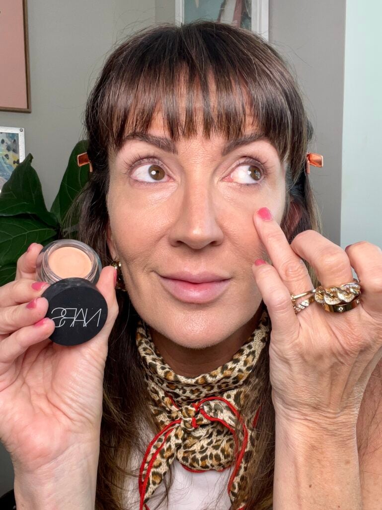 Woman applying Nars Soft Matte Complete Concealer in Creme Brulee under eye with finger, holding open round concealer jar showing nude shade, wearing leopard print neckerchief, gold chain rings, and rose gold hair clips, abstract artwork in background