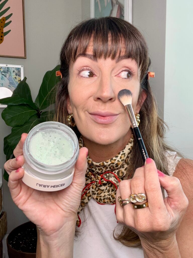 Woman applying Laura Mercier mint green color correcting loose powder with fluffy makeup brush to side of nose, holding open powder jar showing pale green powder, wearing leopard neckerchief and stacked gold rings, fiddle leaf fig plant visible in background