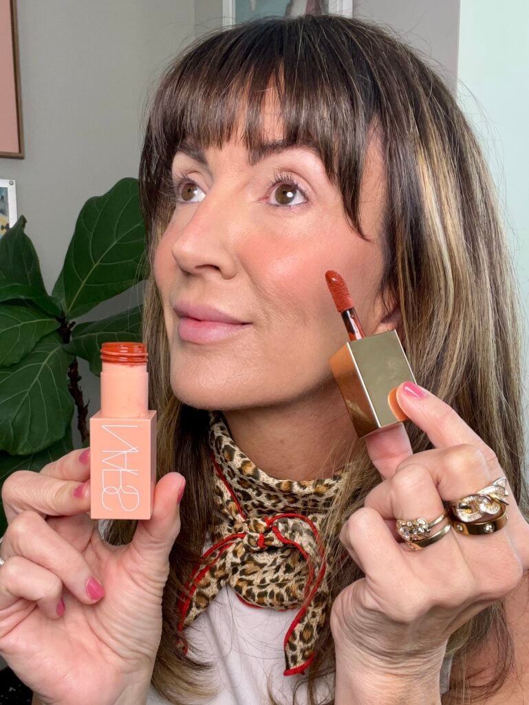 Woman with cream blush flush on cheek, holding NARS Afterglow Liquid Blush in Aragon warm chestnut brown shade with doe-foot applicator and matching peach NARS cream blush stick, wearing leopard neckerchief and stacked gold cocktail rings