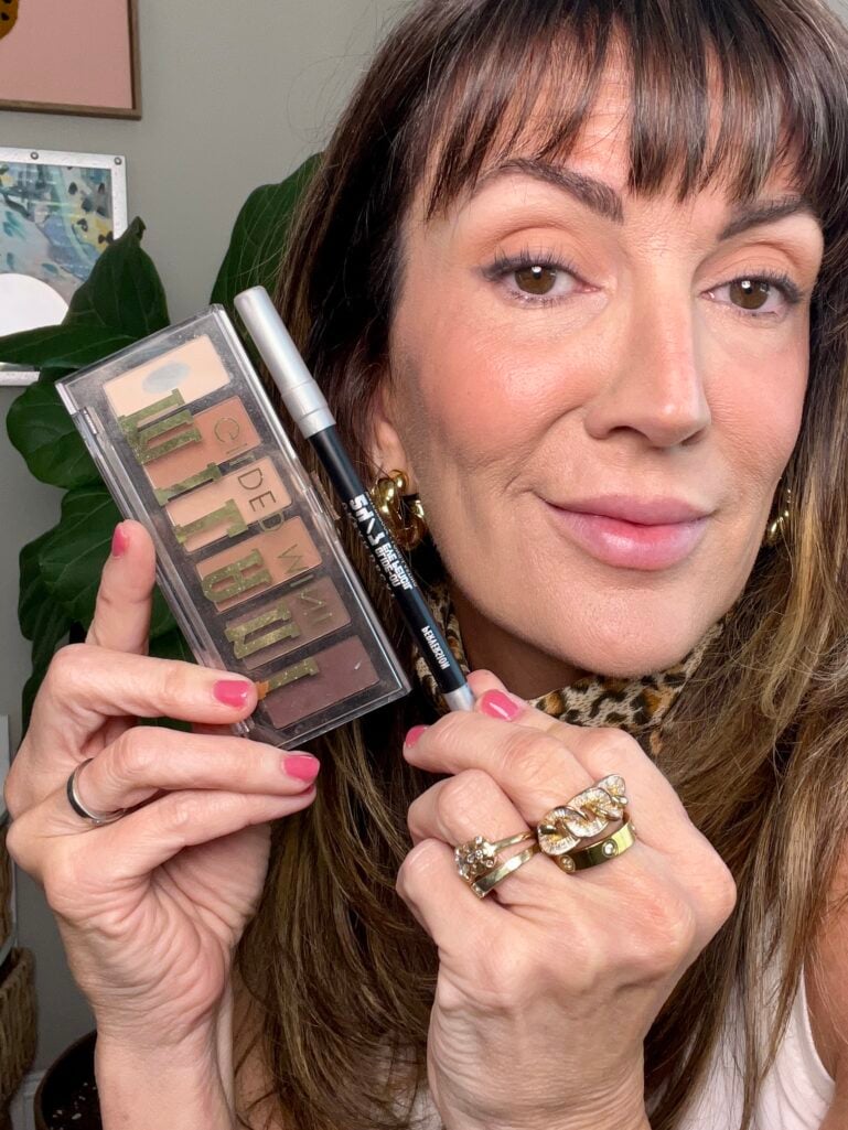 Woman holding Milani Gilded Mini eyeshadow palette in Whiskey Business showing neutral matte browns and Urban Decay 24/7 Glide-On Eye Pencil in Perversion black liner, wearing finished soft brown eye makeup, leopard neckerchief, gold chunky hoop earrings, and stacked gold chain rings