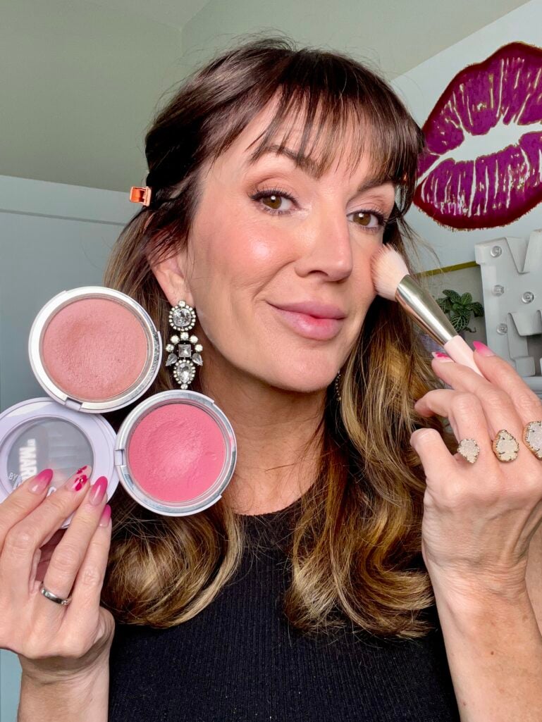Woman applying cream blush with Rare Beauty Soft Pinch brush to high point of cheek, holding Makeup by Mario Soft Pop Plumping cream blushes in Barely Blushing neutral shade and Perfect Pink, wearing crystal earrings and stacked druzy rings