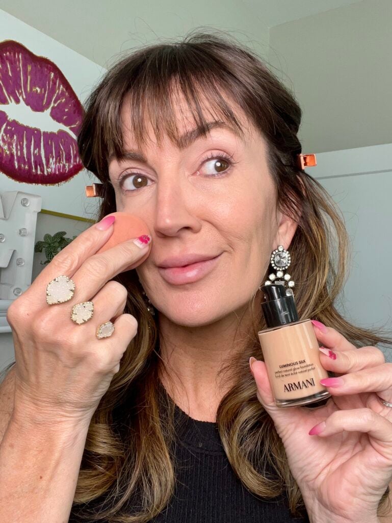 Woman applying Armani Luminous Silk Foundation in shade 4.5 with damp orange Beauty Blender sponge to cheek, holding foundation bottle, wearing crystal earrings, druzy stone Kendra Scott rings, and pink floral nail art with rose gold hair clips