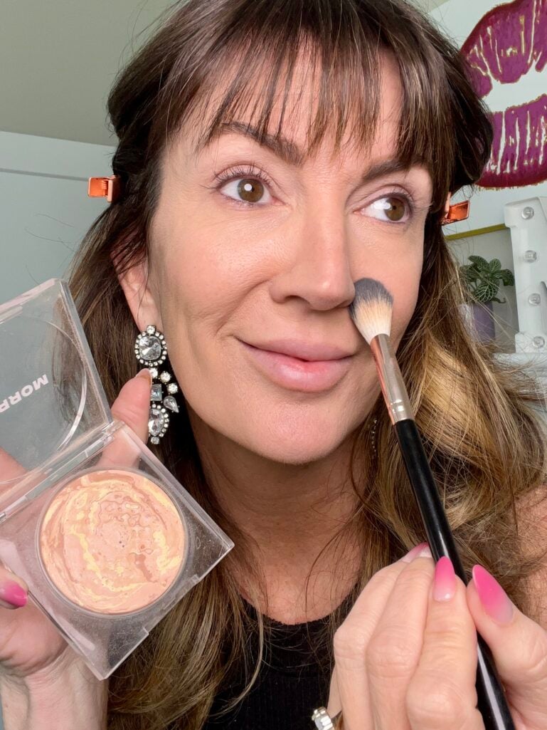 Woman applying Morphe Cloudlight Soft Glow veil powder in shade Sunrise with fluffy makeup brush to side of nose, holding clear powder compact, wearing crystal drop earrings and rose gold hair clips with finished foundation base