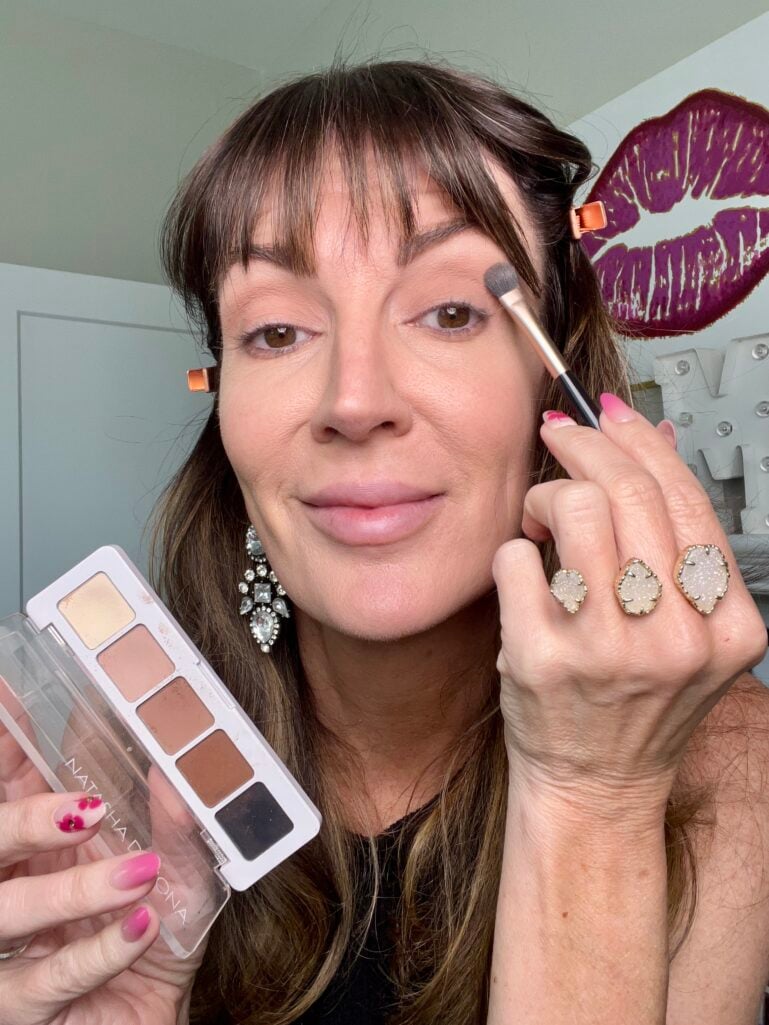Woman applying lighter brown eyeshadow from Natasha Denona Mini Eye Sculpt palette to lid with small fluffy eyeshadow brush, holding palette showing five neutral matte browns, wearing crystal earrings and stacked druzy rings