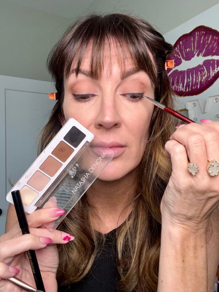 Woman smudging black eyeshadow along upper lash line with tiny BK Beauty precision eyeliner brush, holding Natasha Denona Mini Eye Sculpt palette and Victoria Beckham kajal pencil in cocoa, eyes downcast showing soft smoky brown eye look in progress