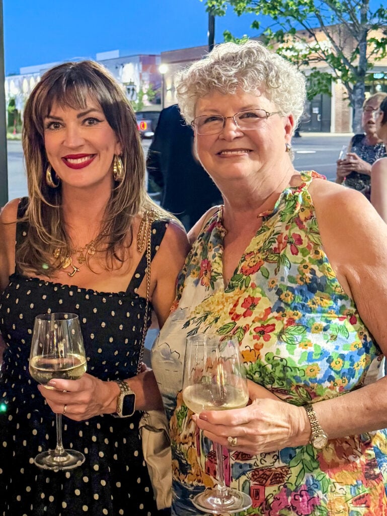 Woman with highlighted brown hair wearing black polka dot sundress with gold layered necklaces and gold hoop earrings holding white wine glass, standing beside smiling older woman with short white curly hair wearing colorful floral sleeveless dress and gold watch, both holding wine glasses at an outdoor evening event with city lights in background