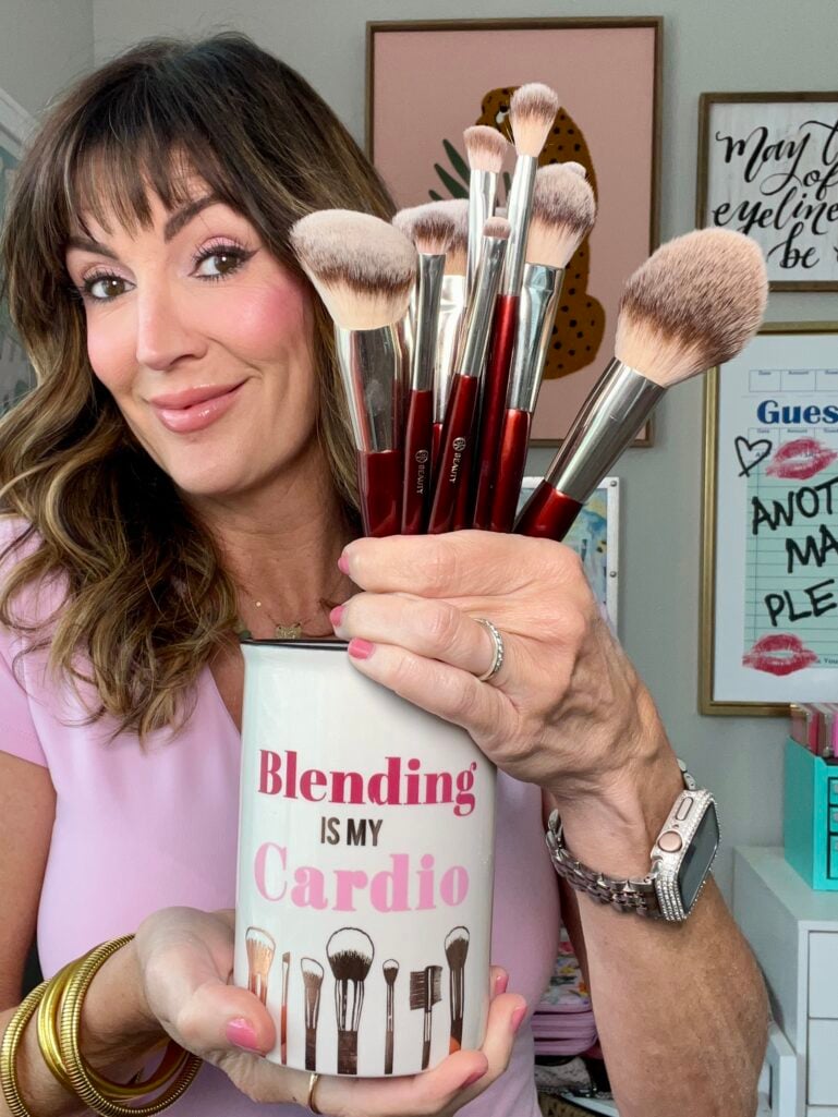 Woman in light pink V-neck top smiling and holding white ceramic "Blending Is My Cardio" makeup brush holder filled with multiple BK Beauty red-handled professional makeup brushes including powder, blush, foundation and eyeshadow styles, wearing gold coil bangles, silver Apple Watch band, pink manicure, gallery wall with cheetah art print and "May Your Eyeliner" and guest book signage in background