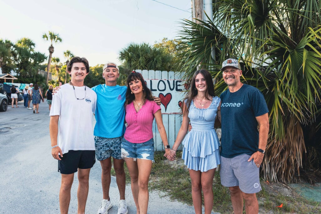 Family of five smiling together on a sunny street near a white painted fence sign reading "LOVE" with a red heart, woman in pink ribbed henley and denim shorts standing between two tall young adult sons in casual summer attire and teenage daughter in light blue smocked dress, man in navy Oconee t-shirt on far right, palm trees and beach town setting in background