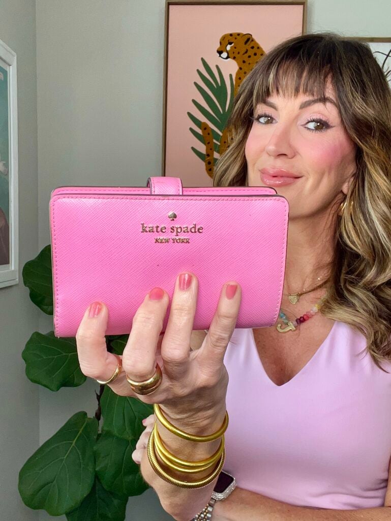 Woman in light pink V-neck top holding up bright pink Kate Spade New York bifold wallet with gold spade logo toward camera while making a playful expression, wearing multicolor beaded necklace, gold dome ring, gold coil bangles and diamond Apple Watch band, fiddle leaf fig plant and leopard cheetah art print in background, pink manicure visible