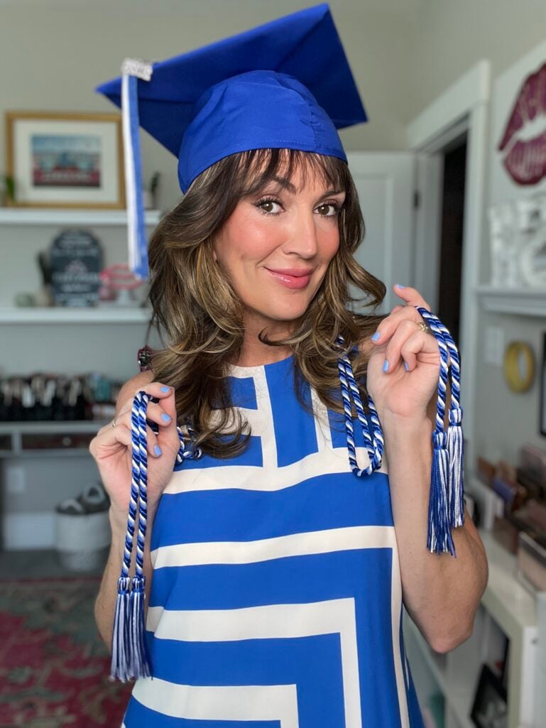 Woman with highlighted brown hair and bangs wearing a blue graduation cap with white tassel and a blue and white geometric print dress, holding blue and white honor cords over her shoulders