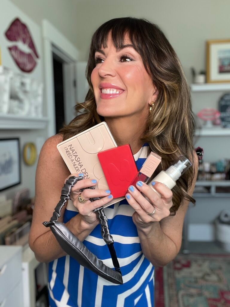 Woman in a blue and white striped dress smiling and looking away while holding an armful of beauty products, including a Natasha Denona nude eyeshadow palette, red blush compact, lip gloss, moisturizer bottle, and a satin sleep mask