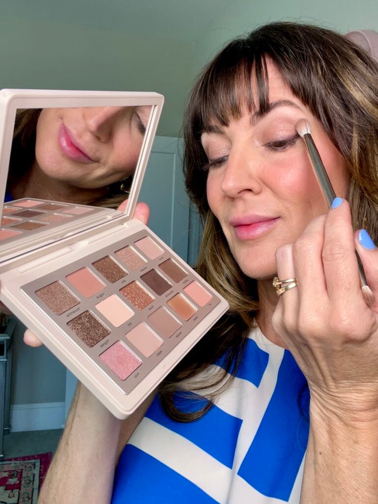 Woman applying shimmery taupe eyeshadow with a brush while holding an open Natasha Denona I Need A Nude palette and handheld mirror, wearing a blue and white dress, with the neutral eyeshadow shades visible