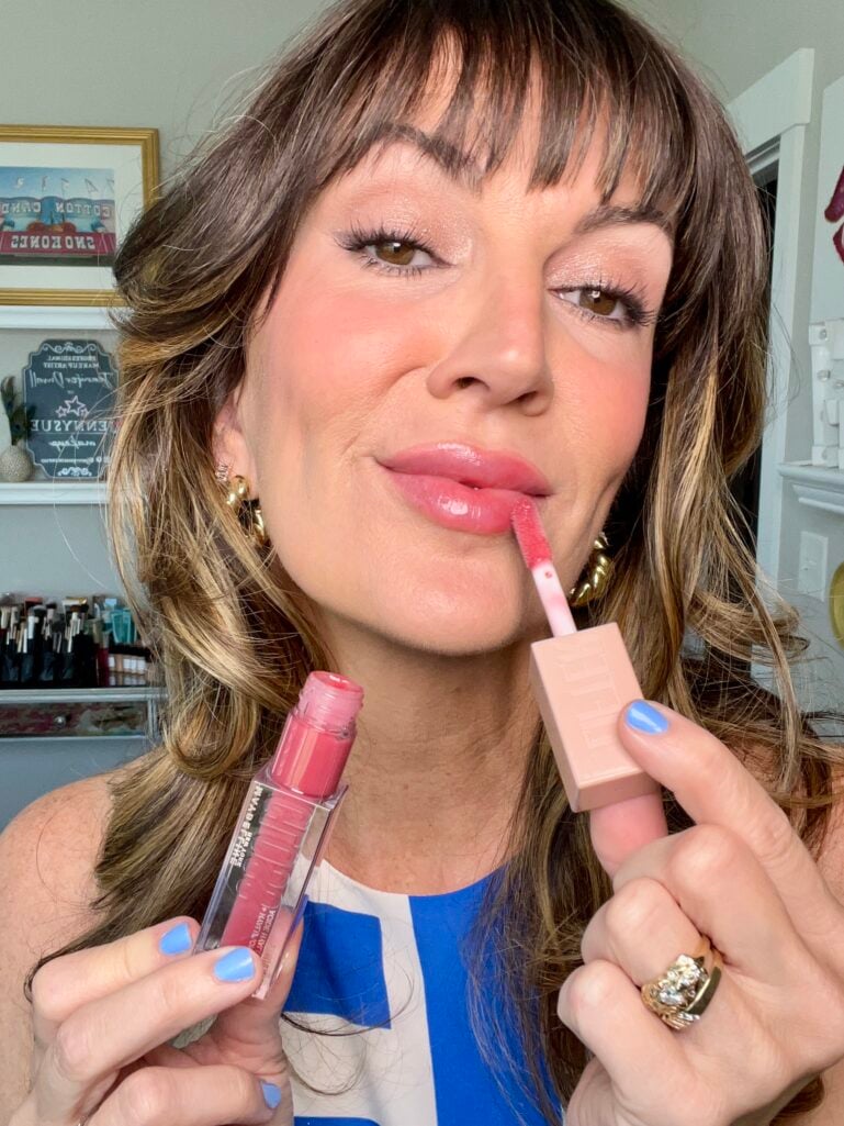 Close-up of a woman applying Maybelline Lifter Gloss in shade Heat to her lips with the doe-foot applicator, holding the pink-tinted tube, wearing soft glam makeup and periwinkle blue nails