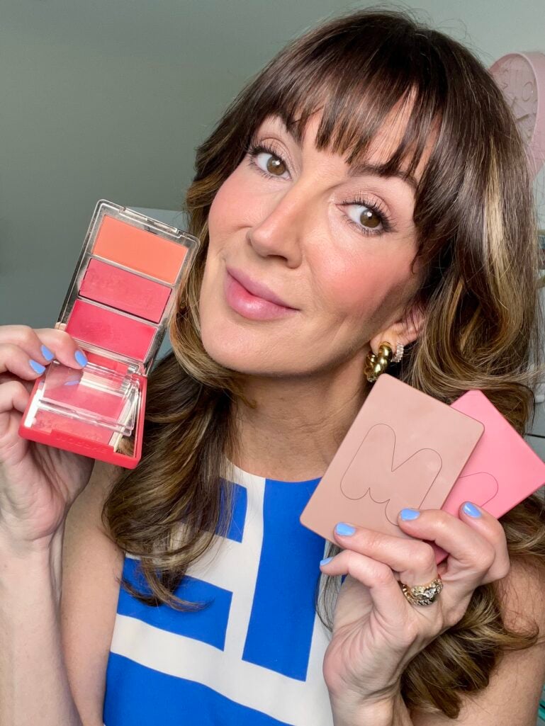 Woman holding an open three-pan coral and red cream blush palette in one hand and Makeup by Mario blush compacts in the other, wearing a blue and white dress, gold earrings, and periwinkle nails with rosy blush on her cheeks