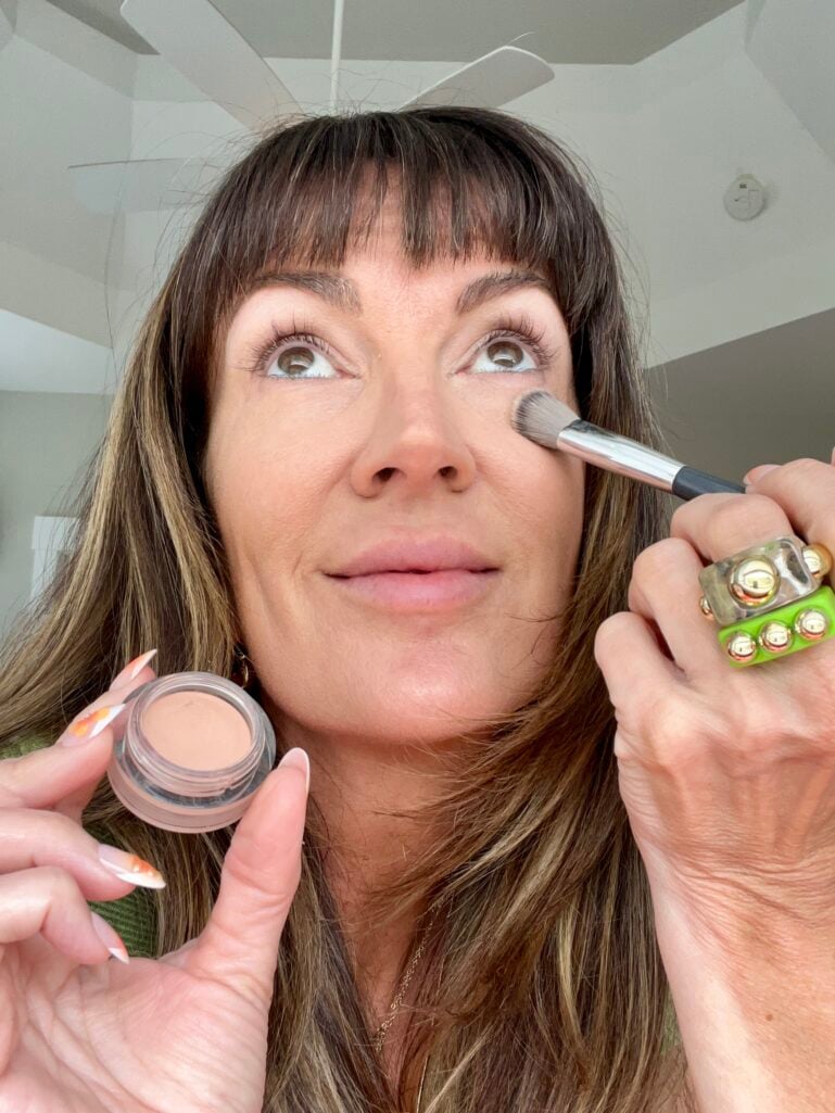 Woman applying Nars Soft Matte Complete Concealer in shade Creme Brulee under her eye with a small fluffy brush, holding the open pot of concealer, wearing chunky gold and green rings