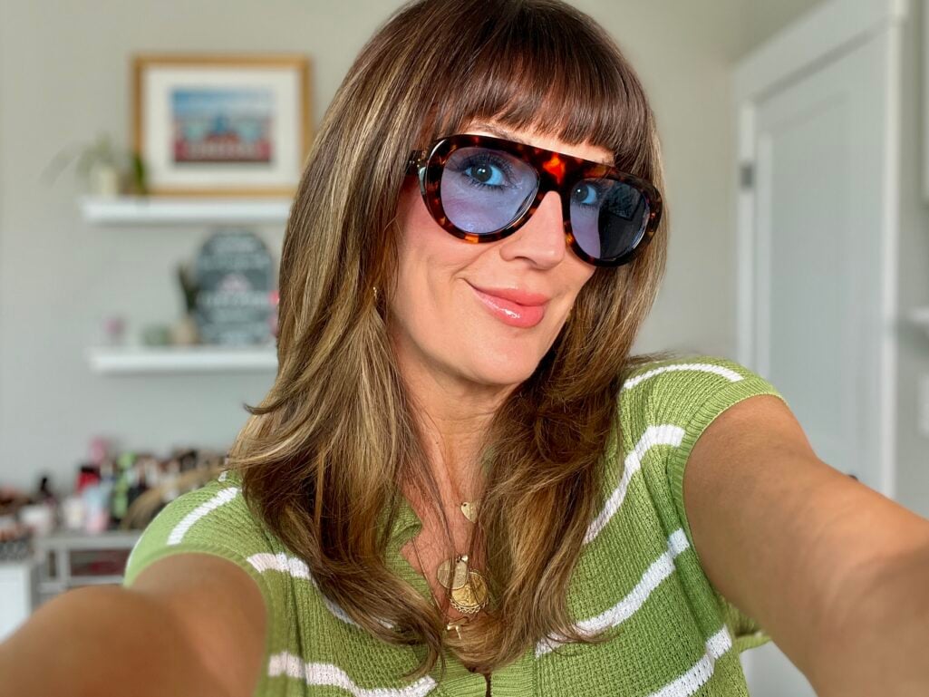 Woman taking a selfie wearing oversized tortoiseshell aviator sunglasses with blue-tinted lenses, glossy nude lips, and a green and white striped knit top