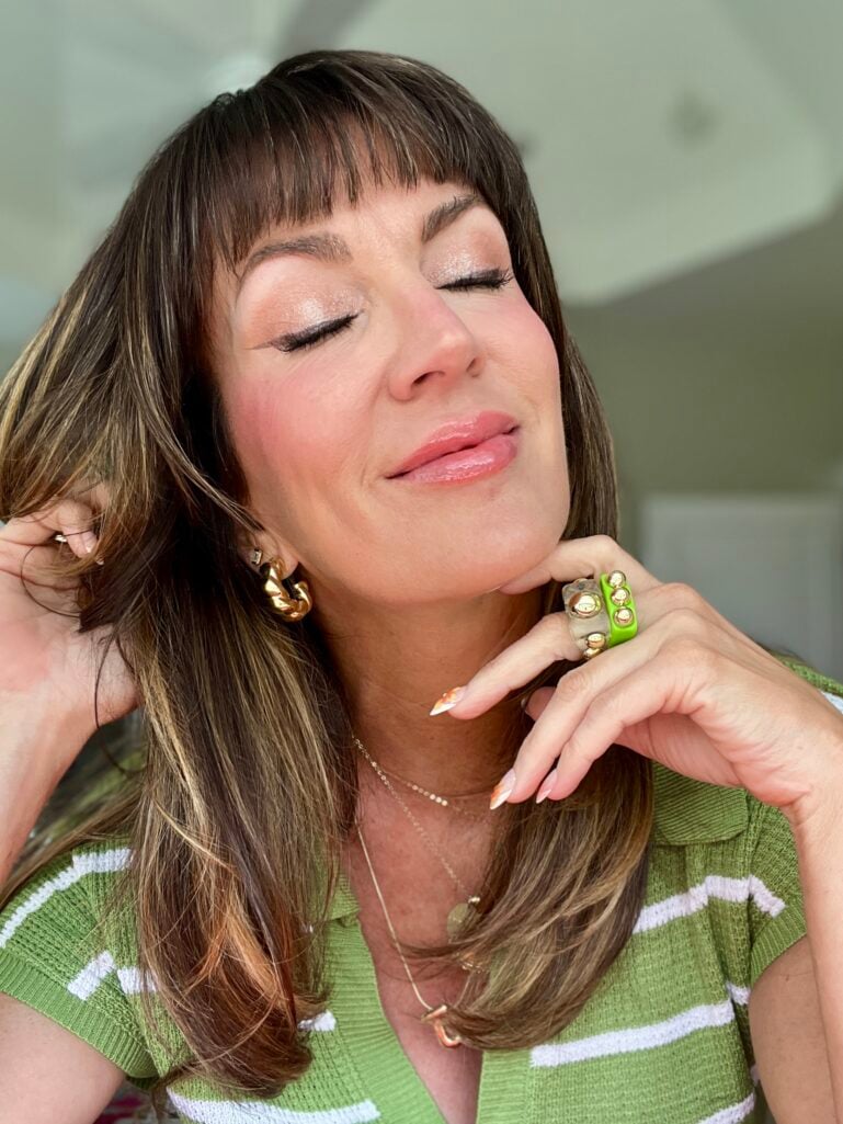 Close-up of a woman with eyes closed showing finished summer makeup look — luminous skin, shimmery champagne eyeshadow, peachy blush, and glossy nude lips, wearing orange floral nail art and stacked gold and green rings