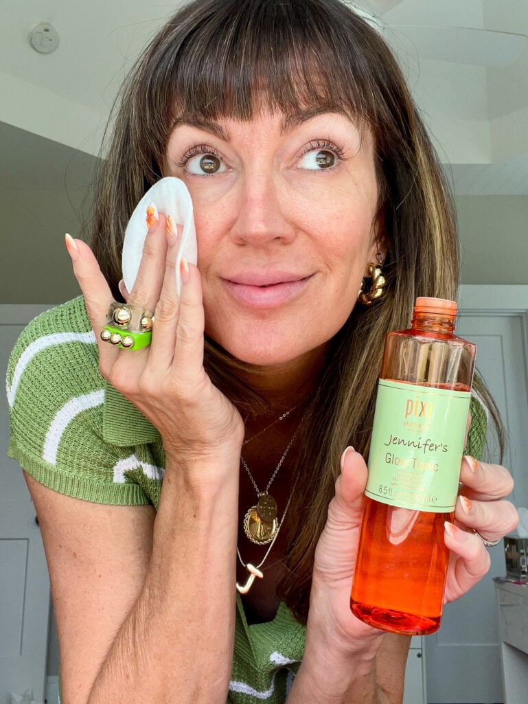 Woman wiping her cheek with a white cotton pad while holding a personalized "Jennifer's Glow Tonic" bottle of Pixi Glycolic Acid exfoliating toner, wearing orange floral nail art and a green striped top