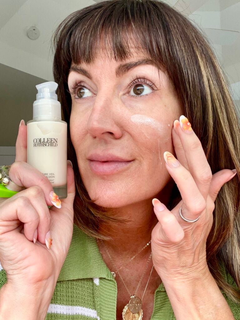 Woman applying Colleen Rothschild Moisture Veil Silk Emulsion to her cheek with her fingertips, holding the frosted glass bottle near her face, showing a fresh dewy finish and orange-and-white floral nail art