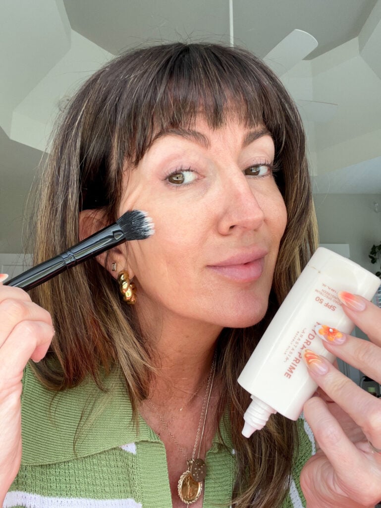 Woman applying Anastasia Beverly Hills Hydra Prime SPF 50 primer to her cheek with a black synthetic-bristle e.l.f. brush while holding the white tube, wearing soft summer makeup and gold earrings