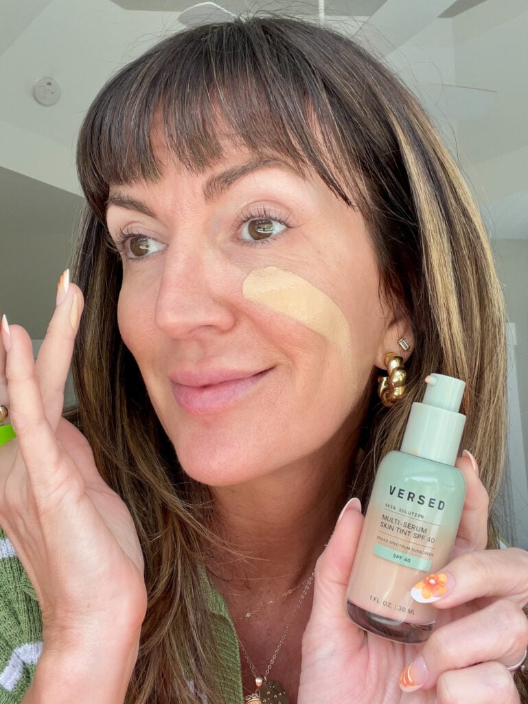 Woman with a swatch of Versed Multi-Serum Skin Tint SPF 40 on her cheek, holding the sage green bottle and showing the lightweight tinted serum coverage before blending