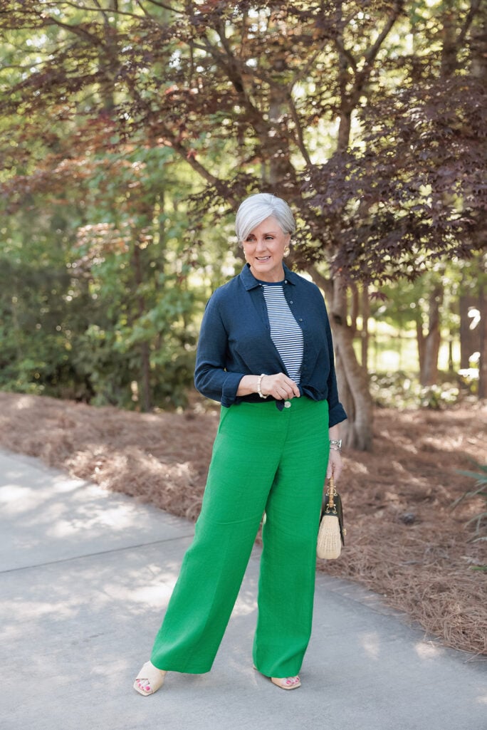 Woman with short silver hair wearing emerald green wide-leg pants, a navy button-front cardigan layered over a navy-and-white striped top, and cream sandals while standing on a sunlit sidewalk. She carries a woven straw handbag with dark leather trim, with lush green trees and a burgundy Japanese maple creating a colorful garden backdrop.