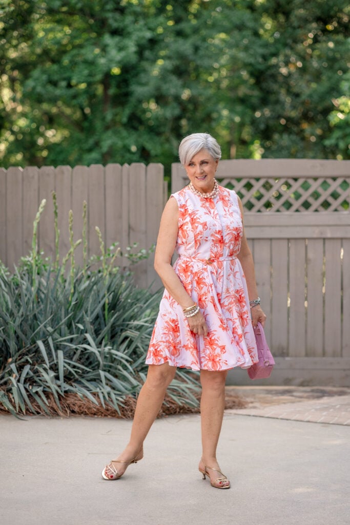 Beth wearing a coral and white floral sleeveless fit-and-flare shirtdress with pearls, gold sandals, and a pink bag
