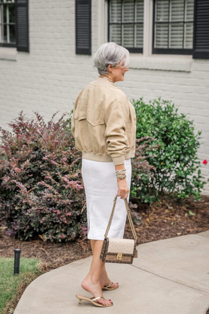 Weekend travel outfit over 50 dressed up — linen bomber jacket, white linen midi skirt, and gold sandals.