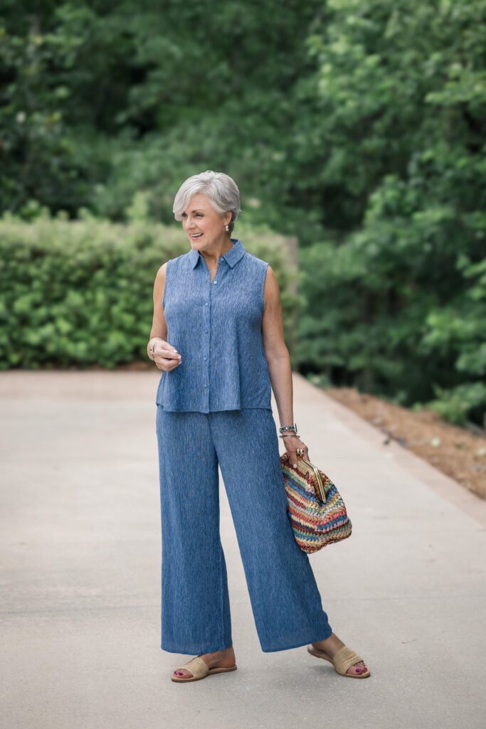Woman over 50 with silver hair walking outdoors in a sleeveless blue crinkle-texture button-up top and matching wide-leg pants from Eileen Fisher, with woven raffia slide sandals and a colorful striped straw bag.