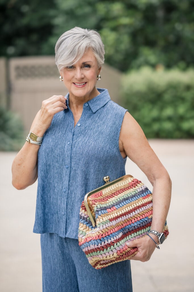 Woman over 50 in a sleeveless blue crinkle-texture top and matching wide-leg pants from Eileen Fisher, turned to the side and adjusting her collar, styled with a striped straw bag.