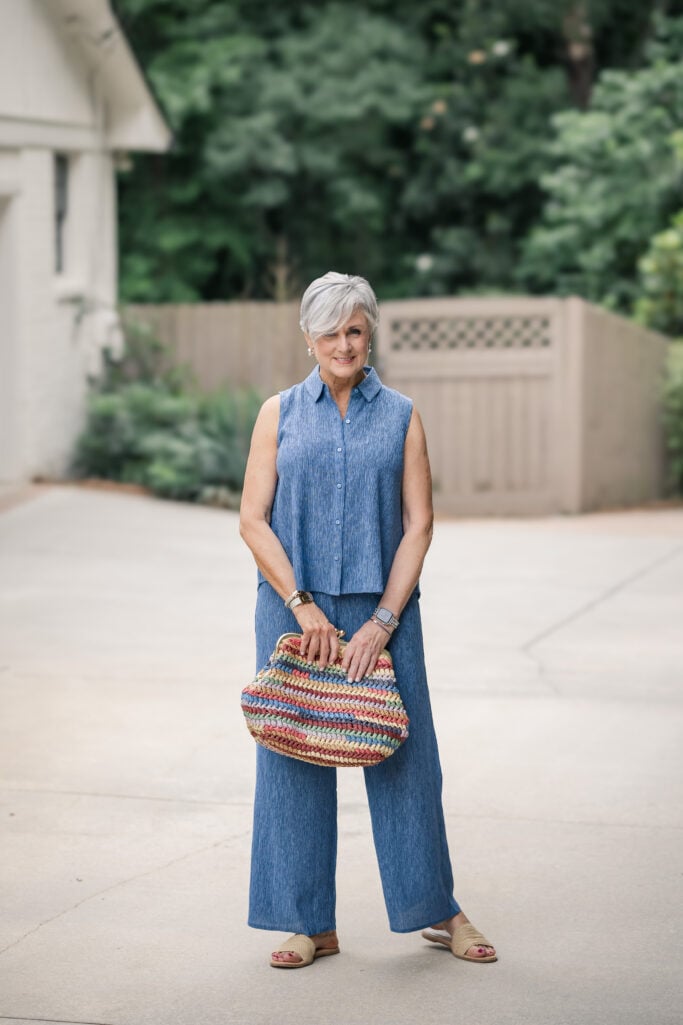 Woman over 50 in a sleeveless blue crinkle-texture top and matching wide-leg pants from Eileen Fisher, styled with a striped straw bag.