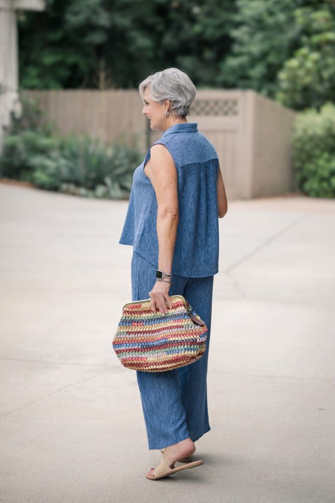 Gray-haired woman standing and holding a multicolor striped raffia clutch with a gold frame closure, wearing a sleeveless blue textured button-up top from Eileen Fisher with a silver watch and stacked bracelets.
