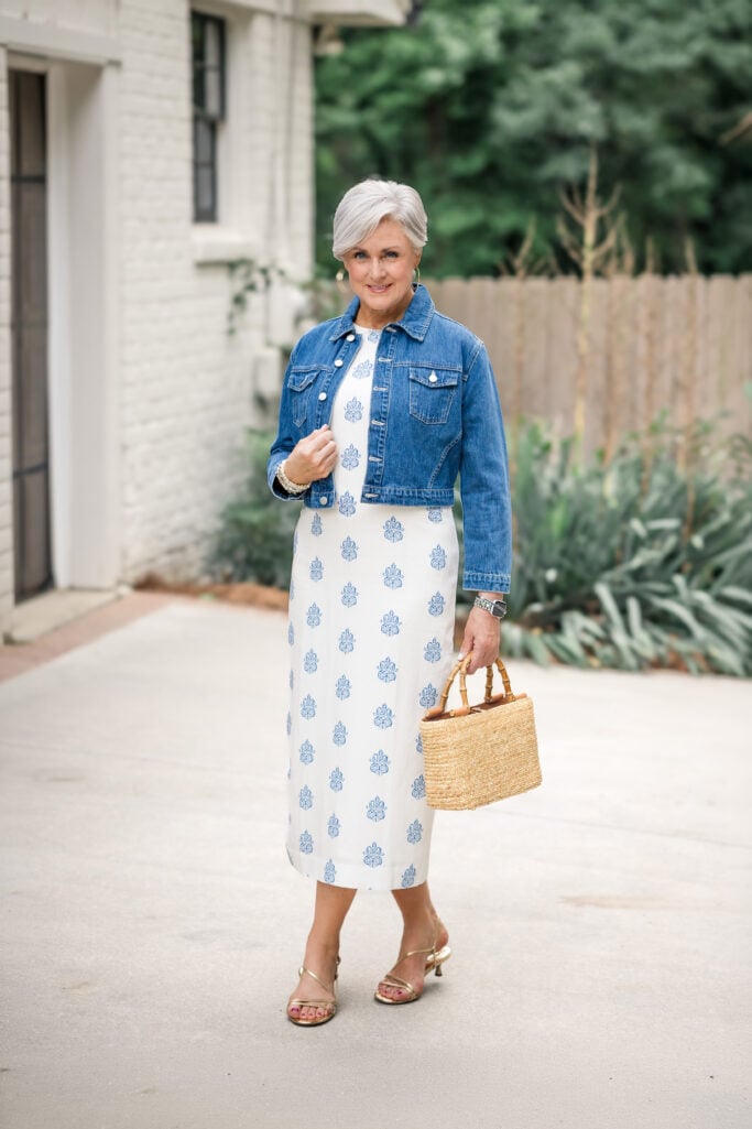 Beth wearing a blue floral midi dress with a cropped denim jacket, metallic sandals, and a woven straw handbag for a summer outfit in Atlanta.