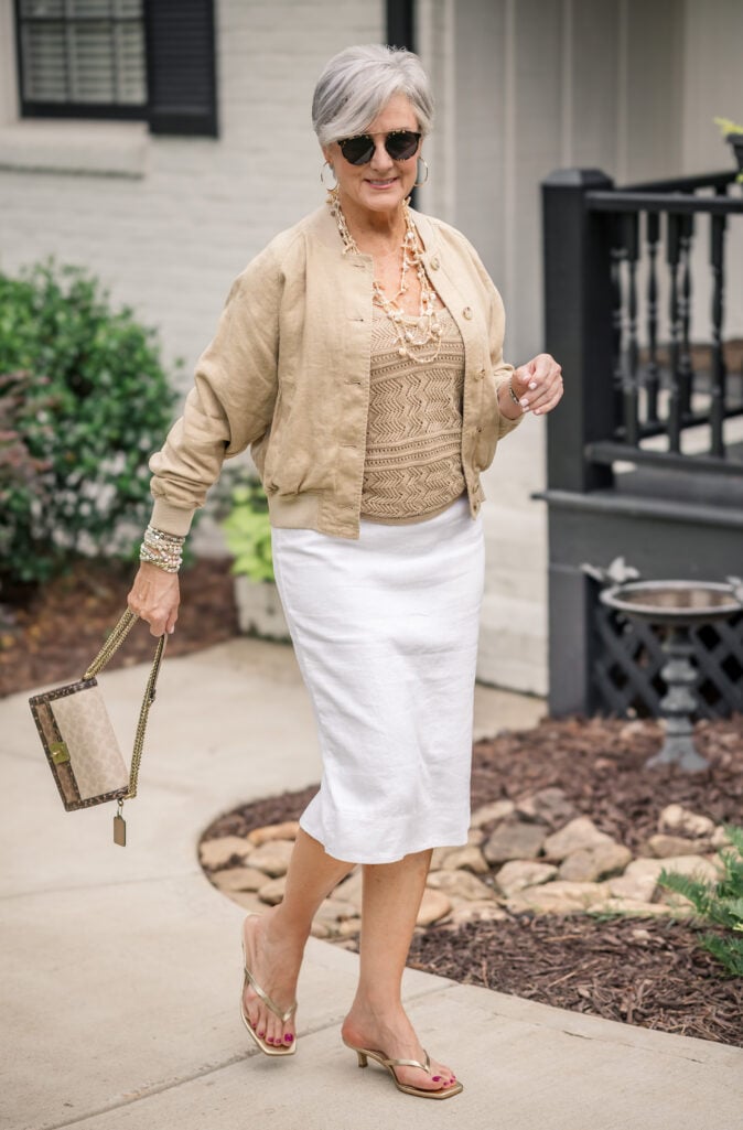 Weekend travel outfit over 50 dressed up — linen bomber jacket, white linen midi skirt, and gold sandals.