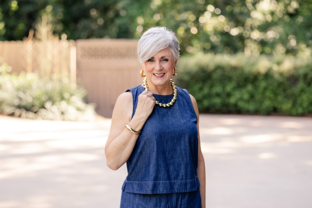 Woman over 50 with short gray hair in a sleeveless boxy denim top and matching wide-leg denim pants from Eileen Fisher, finished with a gold ball-bead necklace, gold hoop earrings, brown leather slide sandals, and a canvas-and-leather tote.