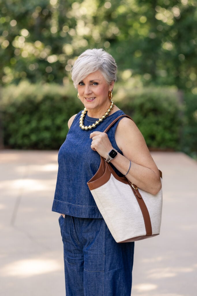 Woman over 50 wearing a sleeveless denim summer outfit with wide-leg pants, leather sandals, and a canvas tote