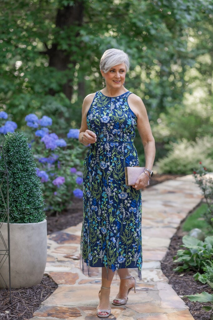 Woman over 50 wearing a blue embroidered midi dress with nude heels for a garden wedding guest outfit.