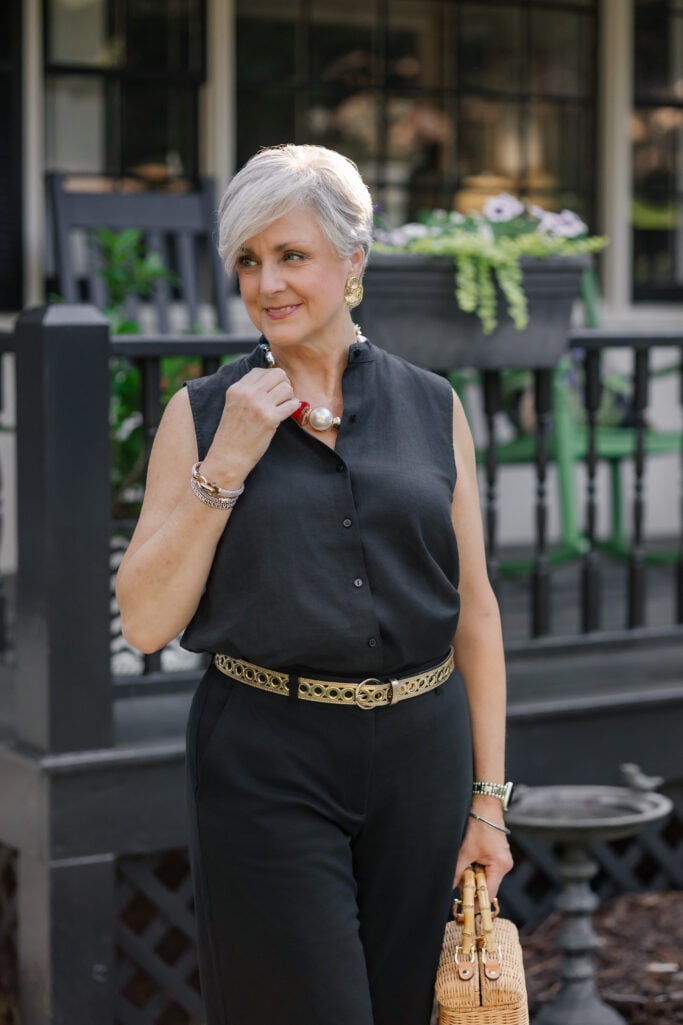Gray-haired woman in a black sleeveless linen top from Eileen Fisher, gold grommet belt at the waist, lifting a chunky pearl-and-coral statement necklace toward her collar.