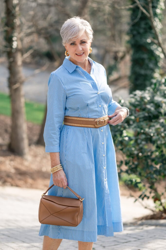 Beth wearing a blue and white striped poplin shirtdress belted at the waist with a woven belt, brown leather bag, and gold hoop earrings — a classic shirtdress style for women over 50
