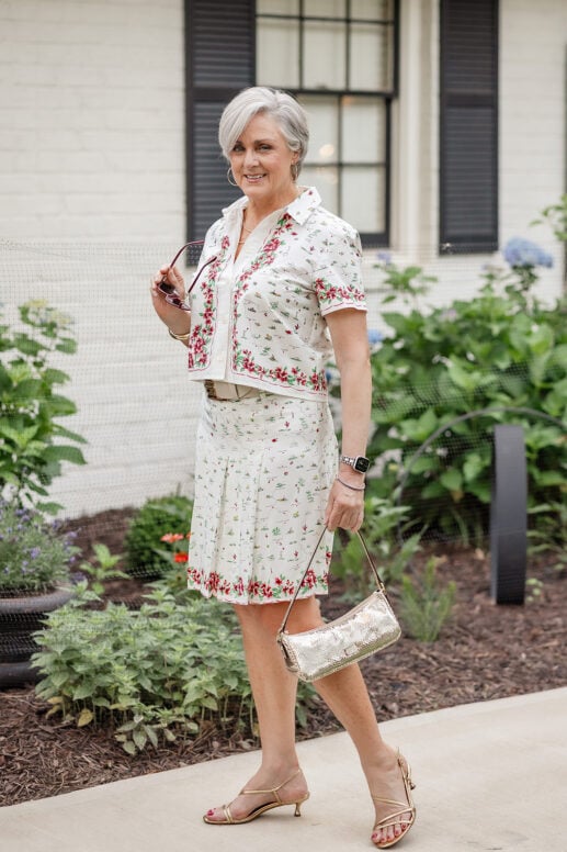 Woman with a silver pixie haircut wearing a coordinated white floral-print blouse and skirt set, gold strappy kitten heels, and a metallic shoulder bag while standing in a lush garden setting outside a white brick home.