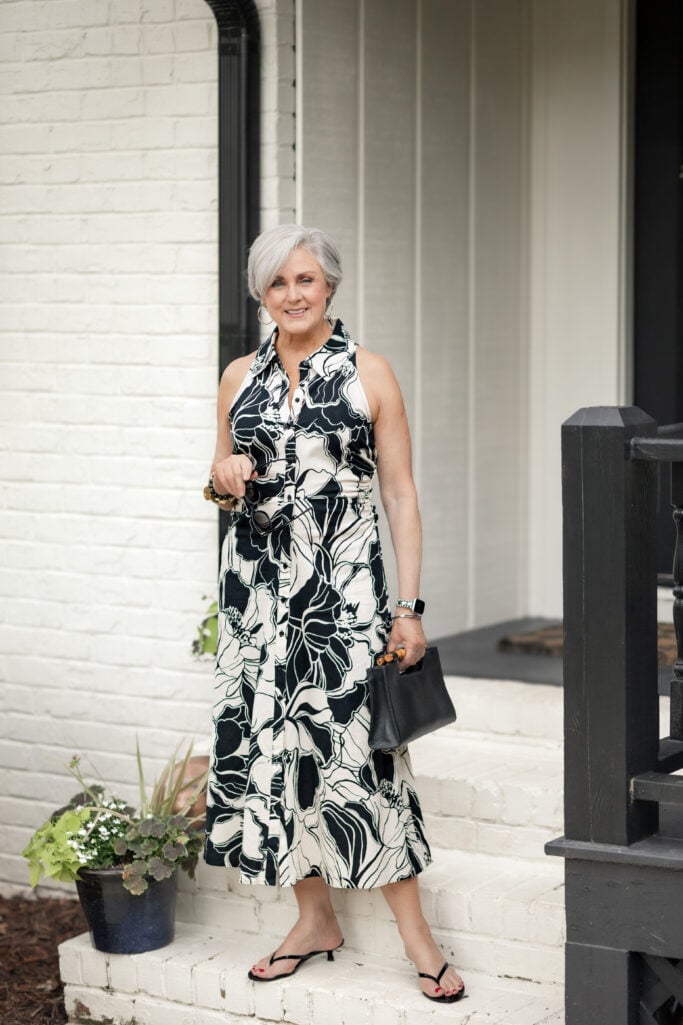Woman over 50 wearing a black and white floral shirtdress with black sandals for a summer wedding guest look.