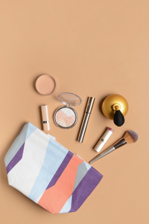 Flat lay of beauty essentials on a beige background, including a striped cosmetic bag, makeup brush, compact powder, highlighter palette, lipstick, mascara, and vintage-style perfume atomizer arranged for travel or everyday use.